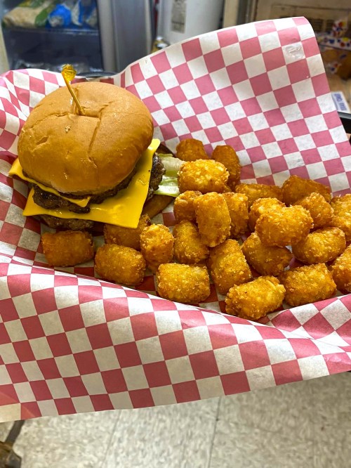 A cheeseburger with a pickle on a bun, served with a side of golden tater tots in a red and white checkered basket.