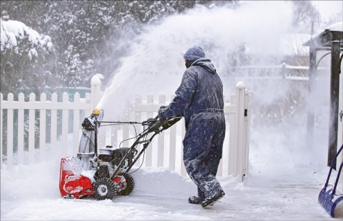 A person in winter gear uses a snowblower to clear snow from a driveway near a white fence.