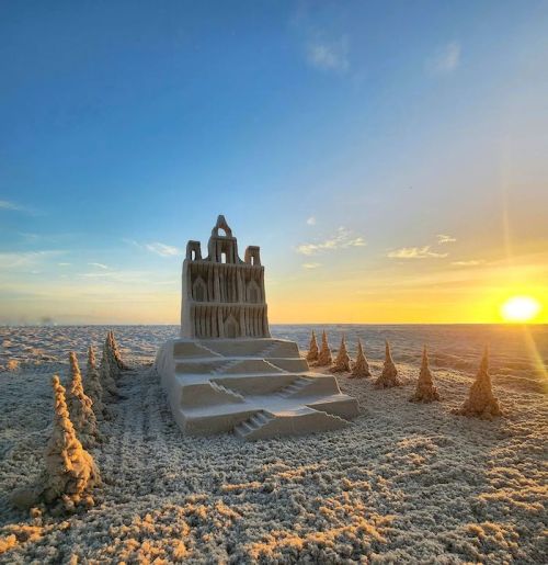 A sandcastle with intricate details stands on a beach at sunset, surrounded by small sand trees.