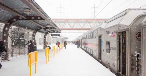 A snowy train station with passengers waiting on the platform as snow falls heavily around them.