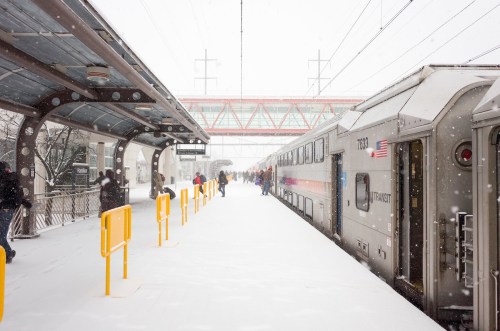 A snowy train station platform with passengers waiting near a train, surrounded by falling snowflakes.