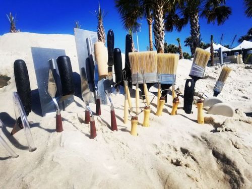 A variety of painting and sculpting tools are partially buried in sandy beach terrain, with palm trees in the background.