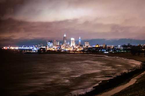 Cleveland skyline at night, with city lights reflecting on the water under a cloudy sky.