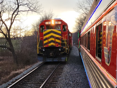 A red and yellow train approaches on the tracks, with a colorful train car visible on the right. Trees line the background.