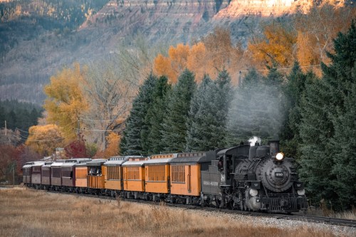 A vintage steam train travels through a colorful autumn landscape with trees and mountains in the background.