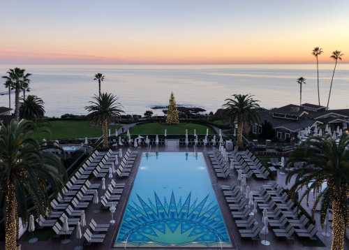 A serene poolside view at sunset, with palm trees, lounge chairs, and a decorated Christmas tree overlooking the ocean.