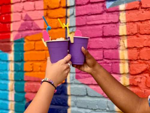 Two hands clink purple cups with colorful straws against a vibrant, painted brick wall.