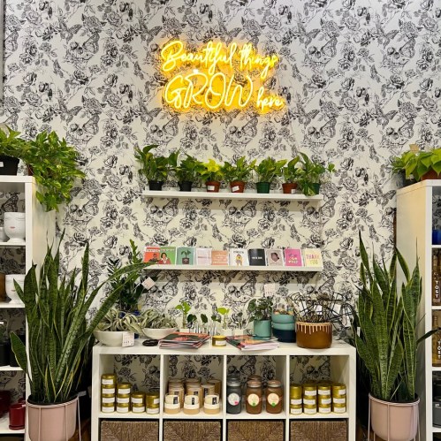 A cozy shop interior with plants, shelves of products, and a neon sign that reads "Beautifully GROWing."