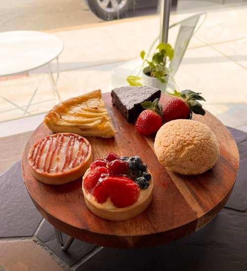 A wooden platter with assorted desserts: fruit tarts, a brownie, a pastry, and two red bean buns.