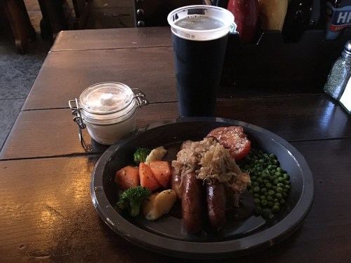 A plate with sausages, sauerkraut, peas, and roasted vegetables, alongside a pint of dark beer and a dessert in a jar.