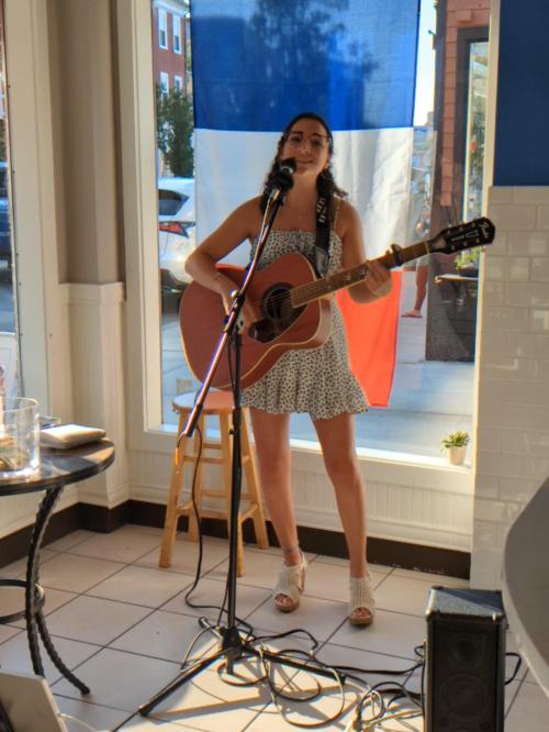 A woman in a polka dot dress sings and plays guitar in front of a large French flag in a cafe setting.