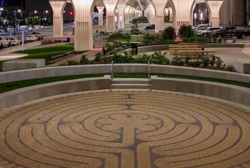 A circular labyrinth design on a stone surface, surrounded by greenery and modern architecture at night.