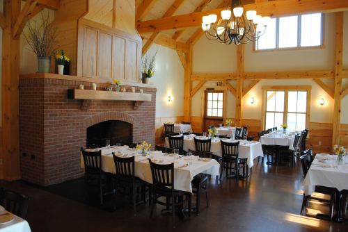 A cozy dining room with wooden beams, a brick fireplace, and tables set with white tablecloths and flowers.