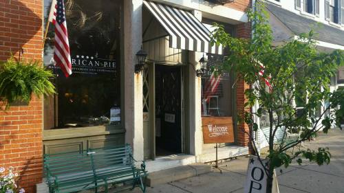 A quaint storefront with a striped awning, flags, and a welcoming sign, surrounded by greenery on a sunny day.