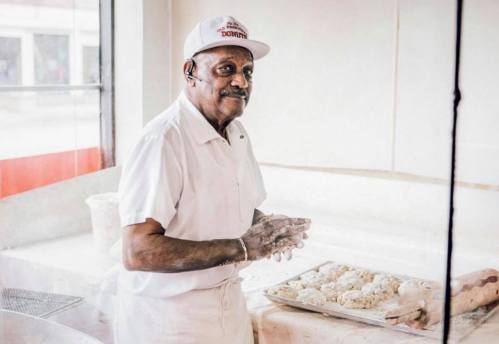 An elderly man in a white uniform and cap prepares dough in a bakery, smiling at the camera.