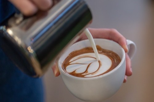 A hand pours steamed milk into a cup of coffee, creating a delicate leaf design in the froth.