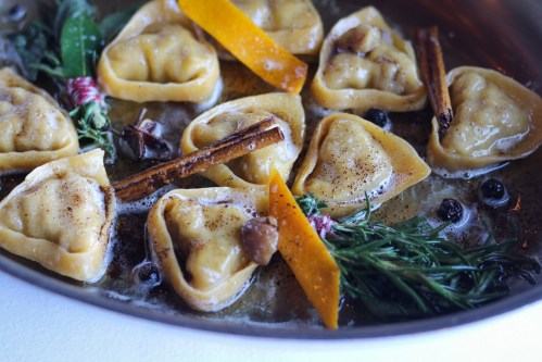A close-up of dumplings in a pan, garnished with herbs, orange peel, and spices, simmering in a flavorful broth.