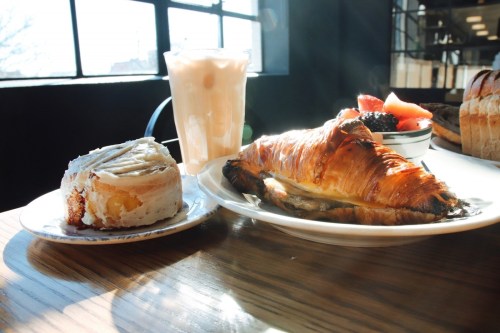A plate with a croissant, a cinnamon roll, and a bowl of mixed berries, alongside a glass of iced drink in a bright café.