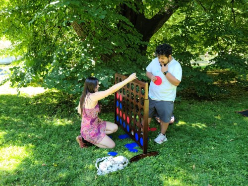 A woman in a pink dress and a man in a white shirt play Connect Four outdoors under a tree.