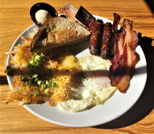 A plate of breakfast featuring eggs, bacon, sausage, hash browns, toast, and condiments on a wooden table.