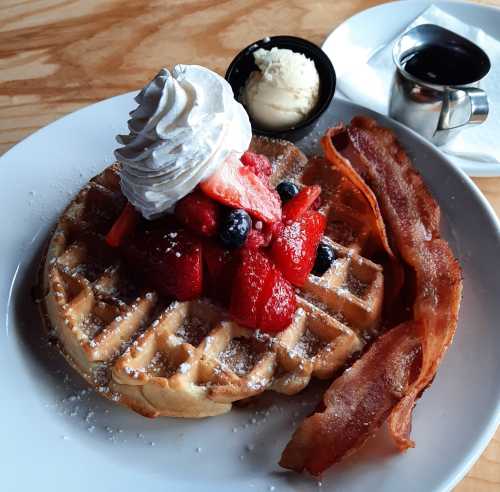 A plate of waffles topped with whipped cream and mixed berries, served with bacon and a side of syrup.