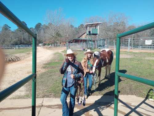 Two people in cowboy hats walk towards the camera, leading a horse in a sunny outdoor setting with a barn in the background.