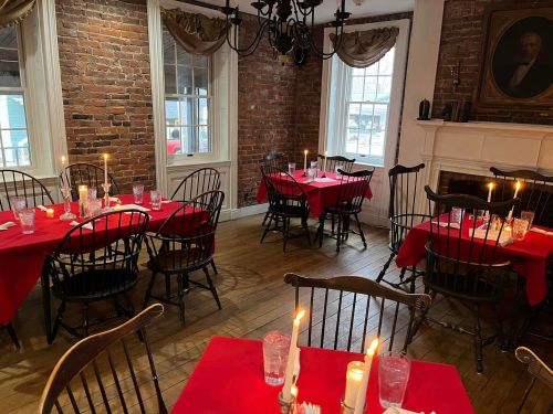 Cozy restaurant interior with red tablecloths, wooden chairs, and candles on tables, featuring exposed brick walls.