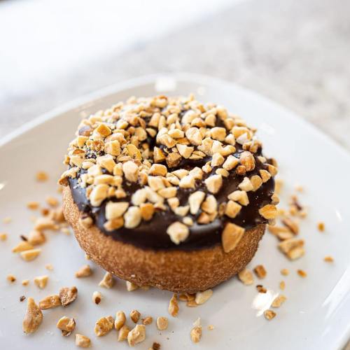 A donut topped with chocolate icing and crushed peanuts, placed on a white plate.