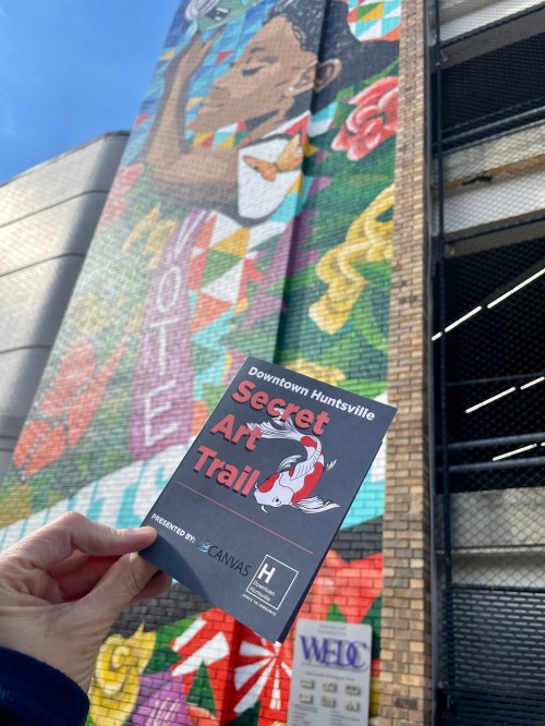 A hand holds a brochure for the Secret Art Trail in front of a colorful mural in Downtown Huntsville.