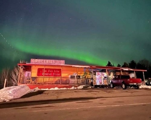 A general store illuminated by the northern lights, with a truck parked outside and snow on the ground.