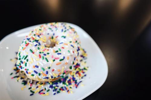 A colorful donut with white icing and sprinkles on a small white plate, surrounded by scattered sprinkles.