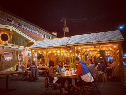 Outdoor dining scene at night with people gathered under illuminated wooden structures, enjoying food and drinks.