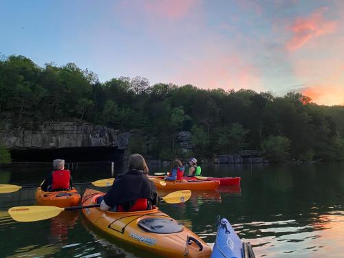 A group of kayakers paddles on a calm lake at sunset, surrounded by lush greenery and rocky cliffs.