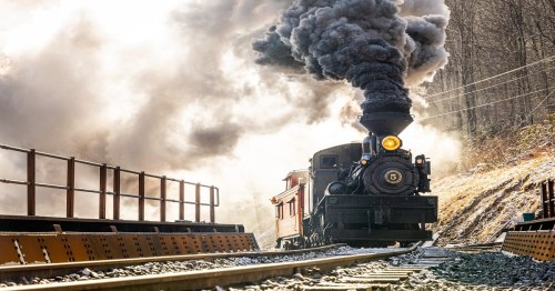A vintage steam train billows black smoke as it travels along a rocky track surrounded by trees.