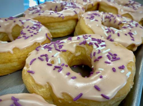 A close-up of glazed donuts topped with pink icing and purple and white sprinkles on a tray.