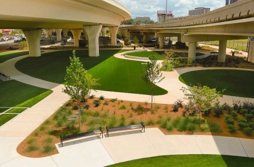 A landscaped park area with green grass, pathways, and benches beneath a highway overpass.