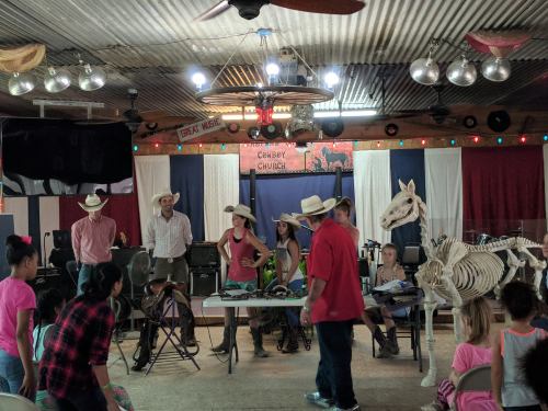 A lively gathering in a barn-like setting with people in cowboy hats, a horse skeleton, and children watching.