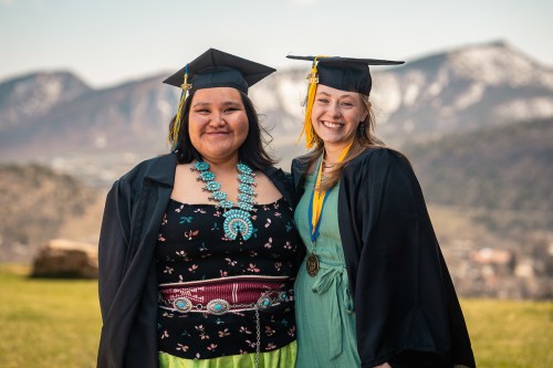 Two smiling graduates in caps and gowns stand together outdoors, with mountains in the background.