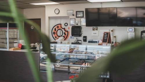 Interior of a bakery with display cases, a clock, and a wall mural, partially obscured by a plant in the foreground.