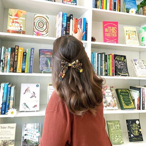 A person with wavy hair and a floral bow stands in front of a bookshelf filled with various colorful books.