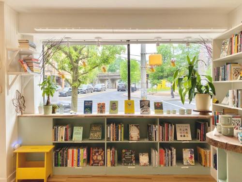 A cozy bookstore window displays shelves of books, plants, and a view of a tree-lined street outside.