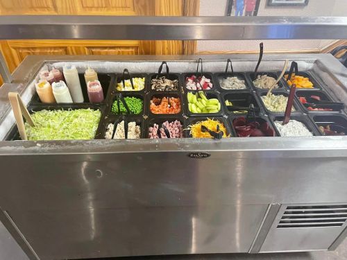 A salad bar featuring various toppings, including vegetables, sauces, and proteins, arranged in labeled containers.