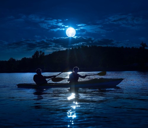 Two people kayak on a calm lake under a bright full moon, surrounded by dark clouds and trees.