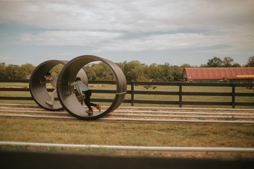 Two children play inside large metal tubes on a grassy field, with a red barn and trees in the background.