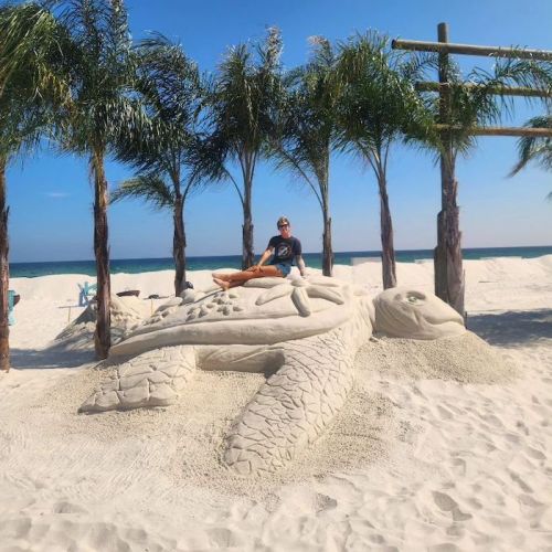 A person sits on a large sand sculpture of a turtle, surrounded by palm trees and a beach backdrop.