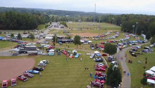 Aerial view of a car show with numerous vehicles and tents set up in a grassy area, surrounded by trees and fields.