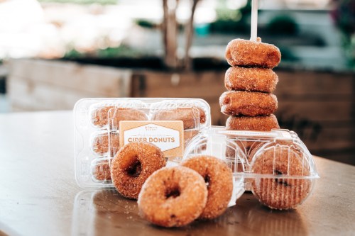 A stack of cider donuts next to a clear container filled with more donuts, set on a wooden table.