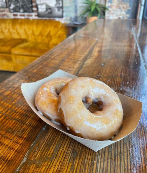 Two glazed donuts on a paper tray, resting on a wooden table with a cozy background.