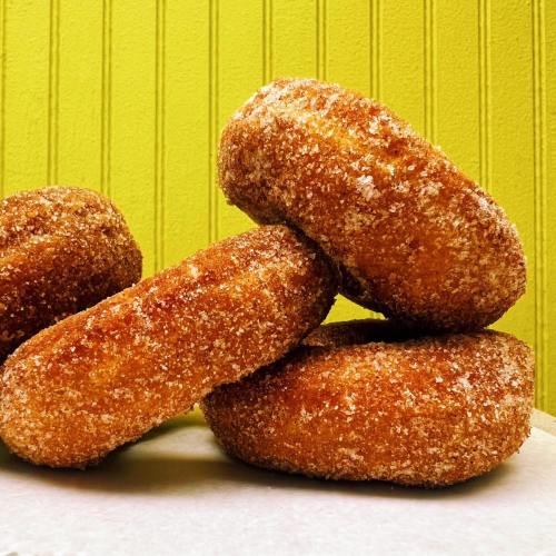 Four golden-brown, sugar-coated donuts stacked on a white surface against a bright green background.