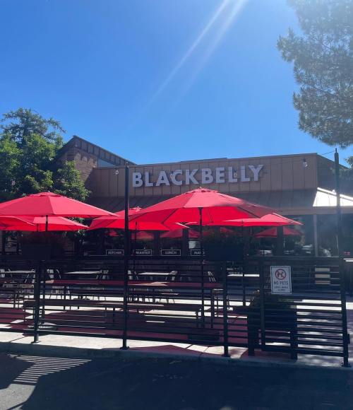 Exterior of Blackbelly restaurant with red umbrellas and a clear blue sky. No pets allowed sign visible.